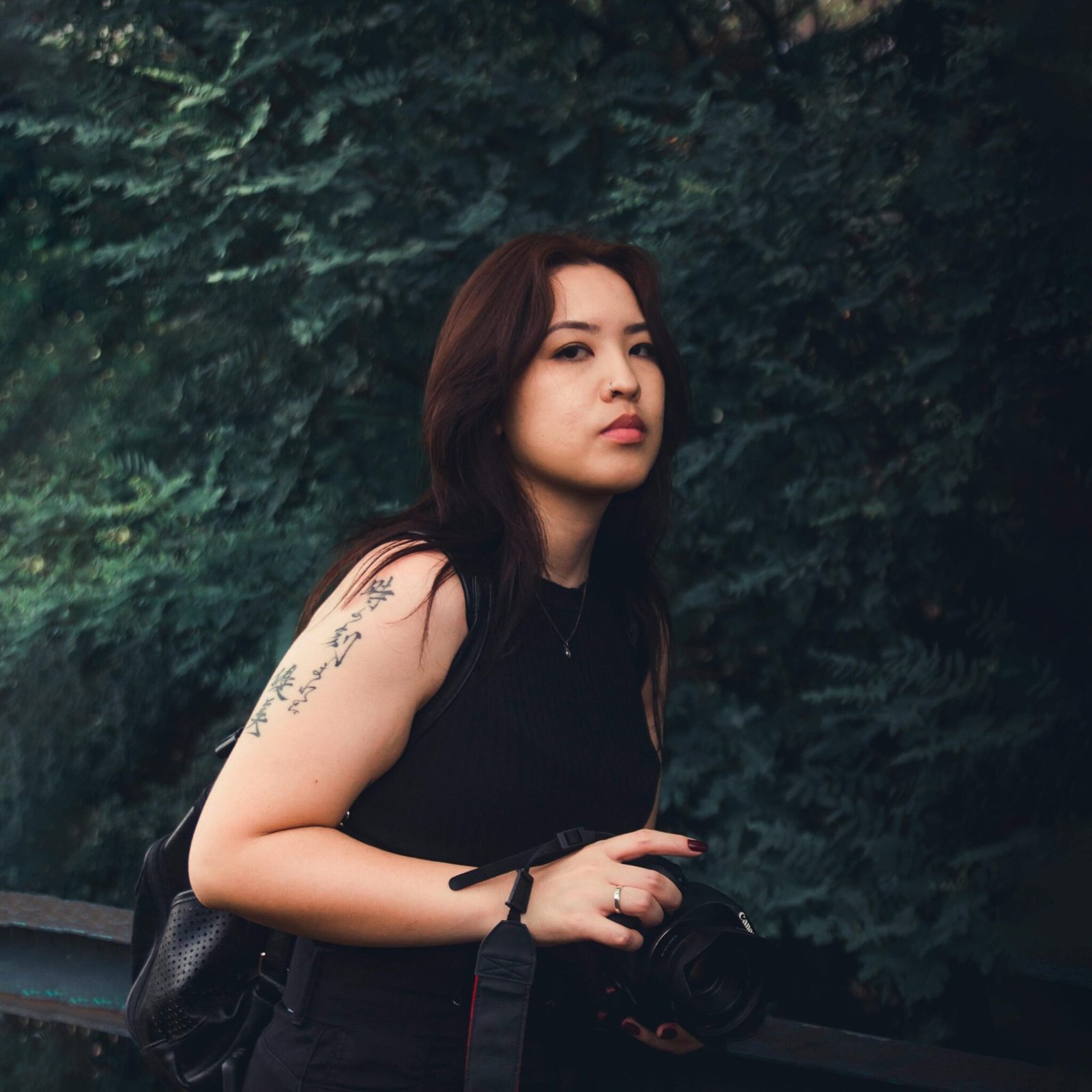 Portrait of a young woman with a tattoo posing elegantly outdoors against lush greenery.